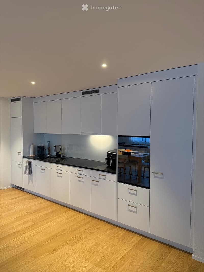 Modern kitchen with white cabinets, black countertops, and a reflective backsplash. Visible items include a sink, faucet, coffee maker, and oven. A small dining area with a table and chairs is visible through the backsplash.