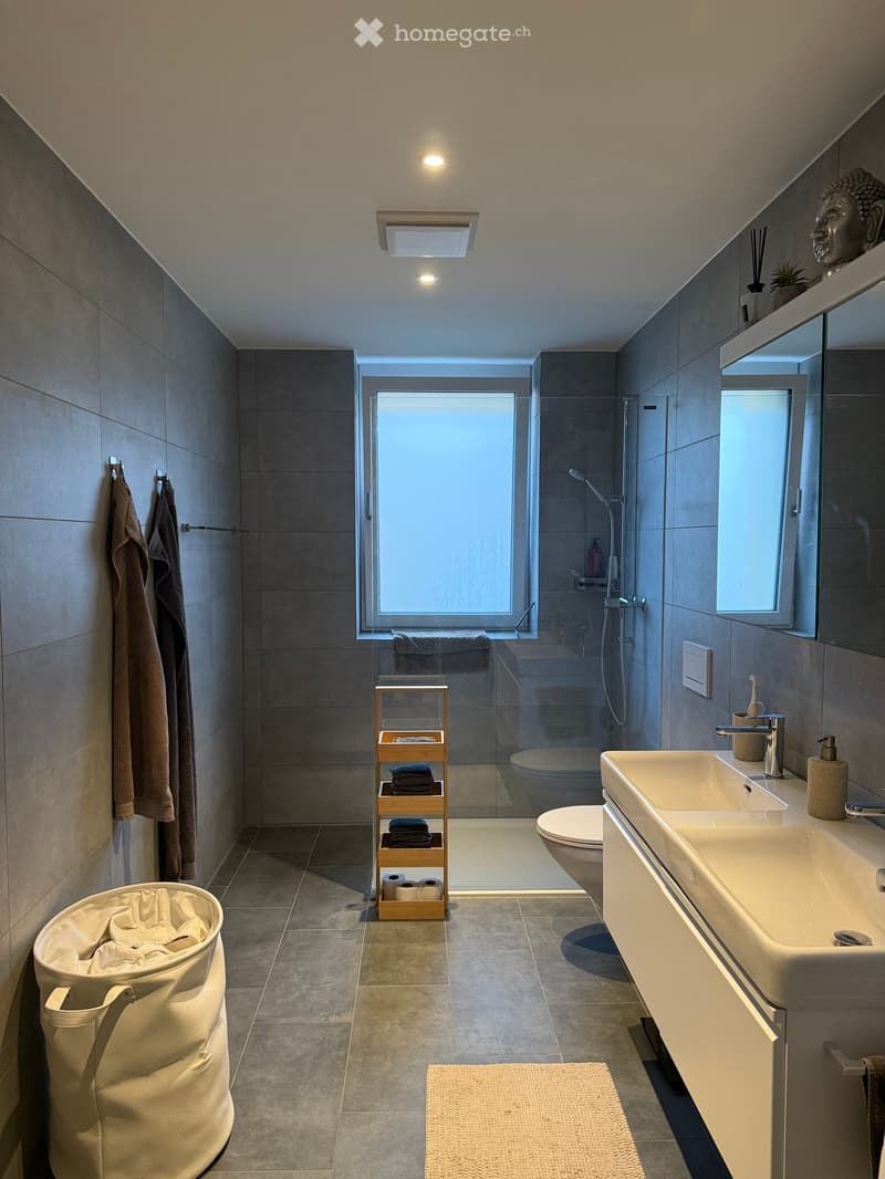 Modern bathroom with double sink, toilet, shower, towel rack, and wooden shelf holding towels and toilet paper. Features a large window, sleek fixtures, and a neutral color palette.