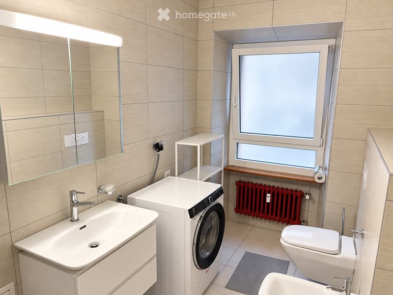Modern bathroom with a white sink, toilet, washing machine, and a large mirror above the sink. The room features light wooden tiles and a red radiator under the window.