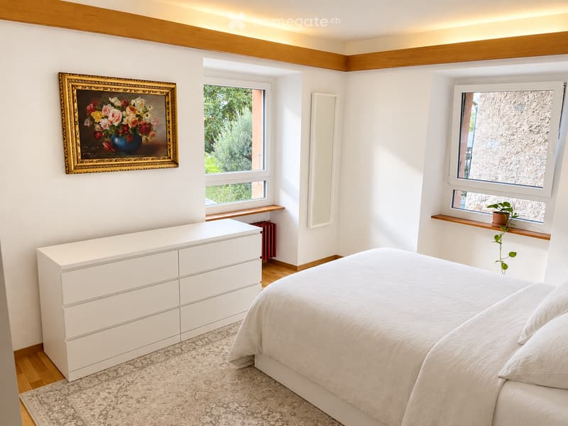 Modern bedroom with a large bed, white dresser, framed painting, small plant on windowsill, and radiator.