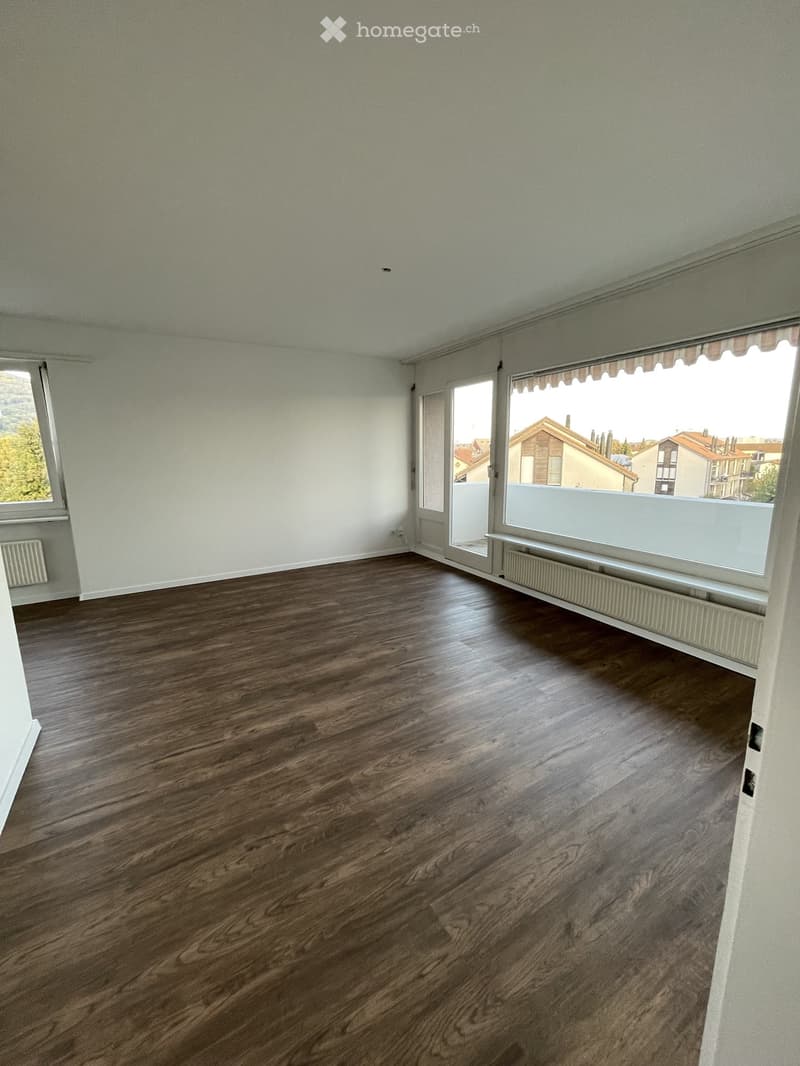 Empty room with wooden flooring, white walls, large windows, and a sliding glass door leading to a balcony.
