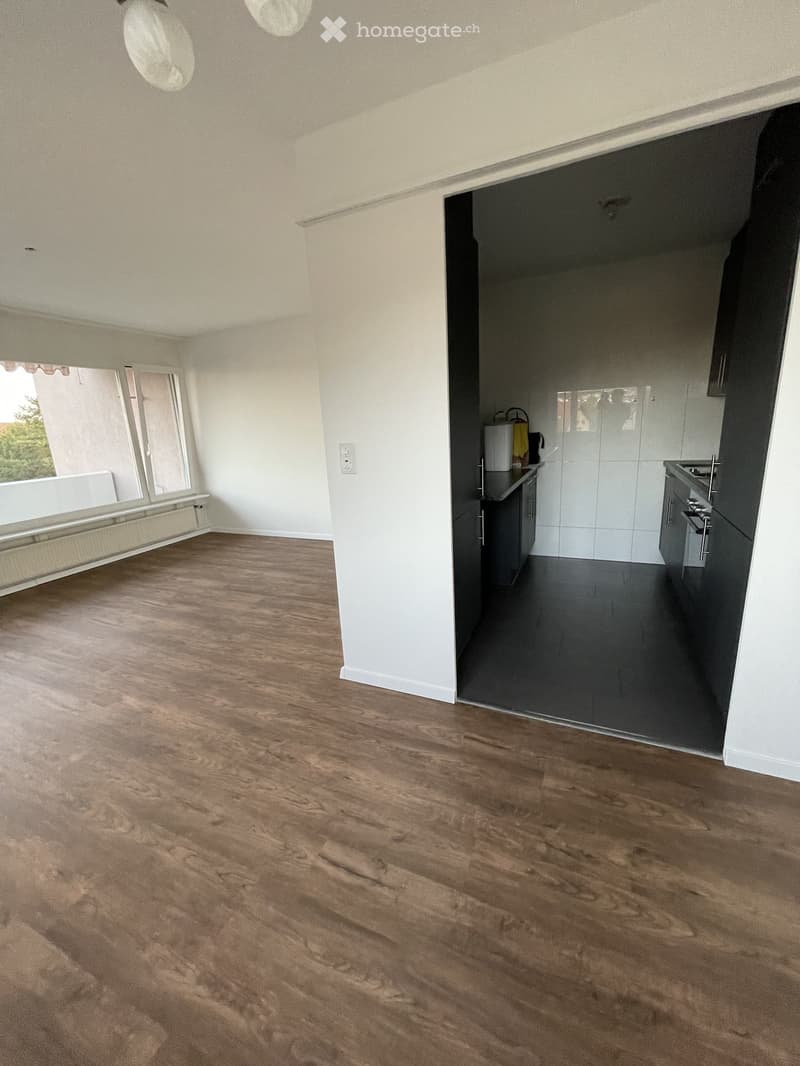 Empty room with wooden flooring, white walls, and a view of a kitchen area with cabinets and appliances.