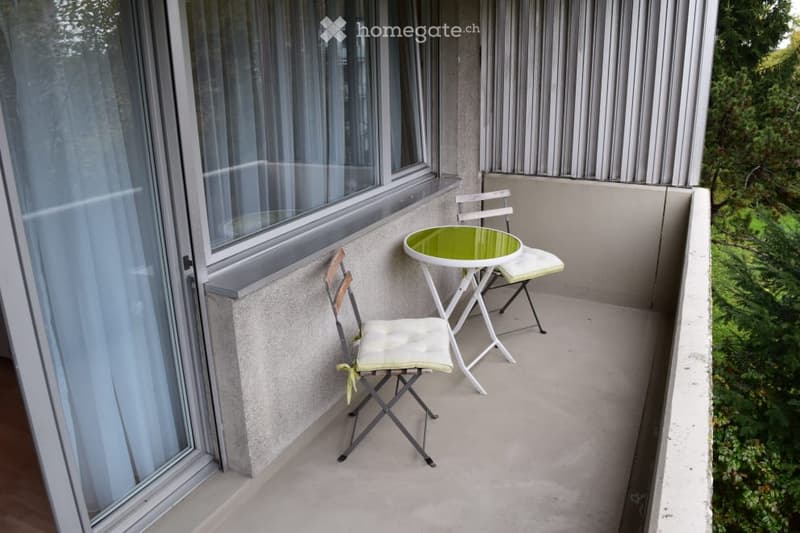 Balcony with a small round table and two folding chairs, large windows with curtains, and a view of greenery.