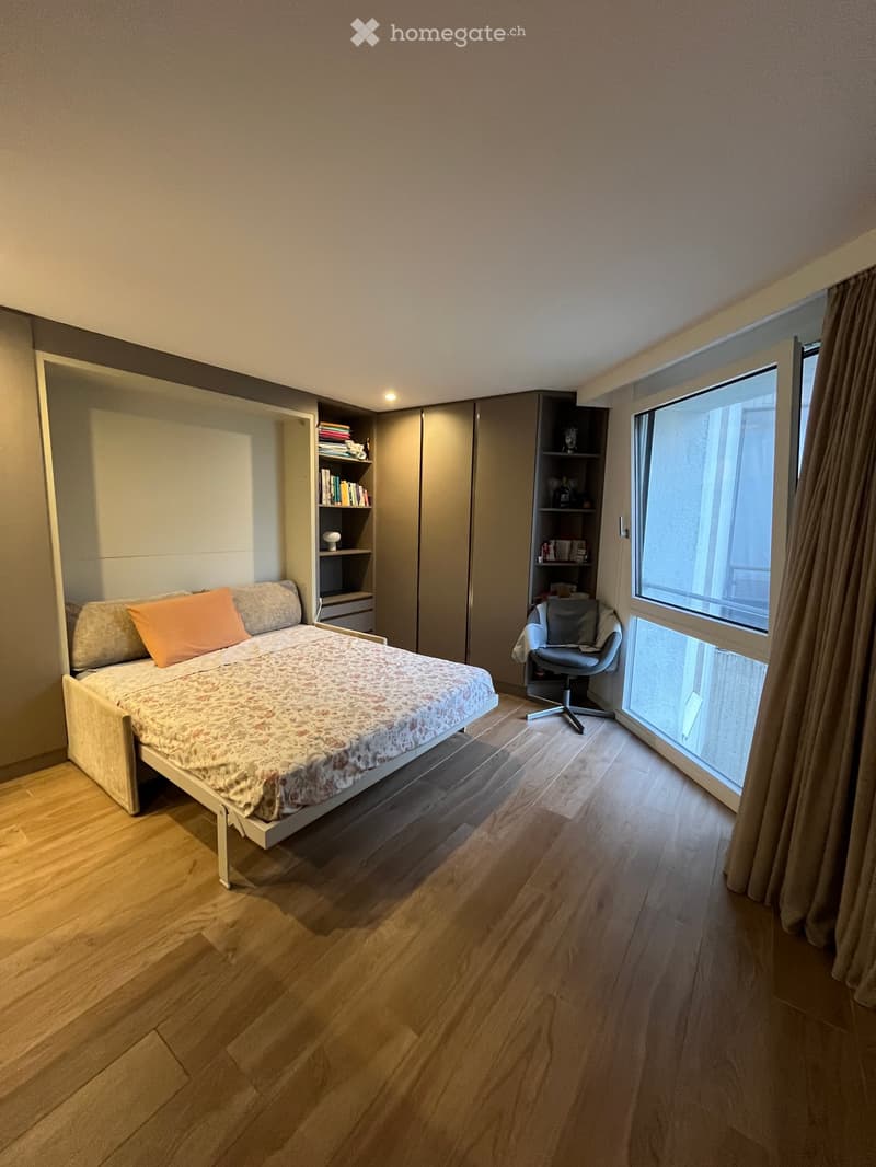 Modern bedroom with a single bed, a chair, a wardrobe, and a bookshelf. The room features wooden flooring and large windows with curtains.