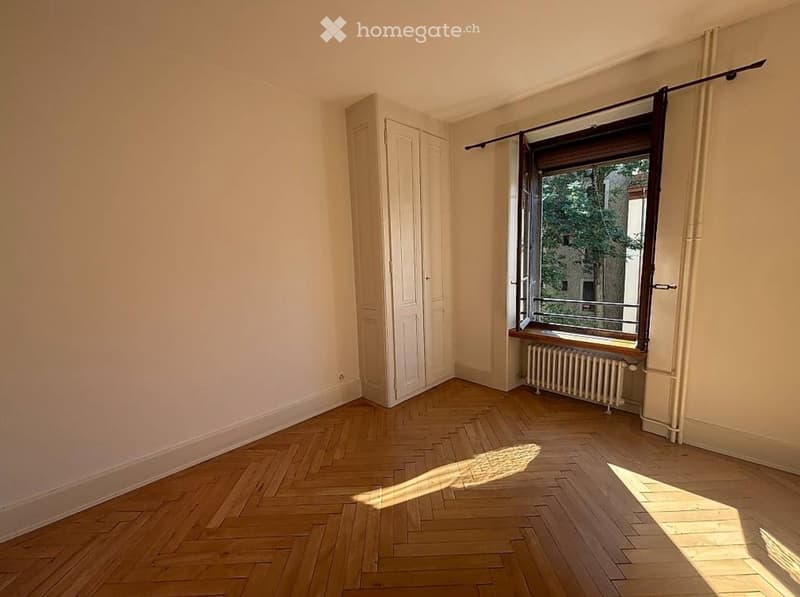 Empty room with wooden herringbone flooring, white walls, a window with a radiator below, and a closet with double doors.