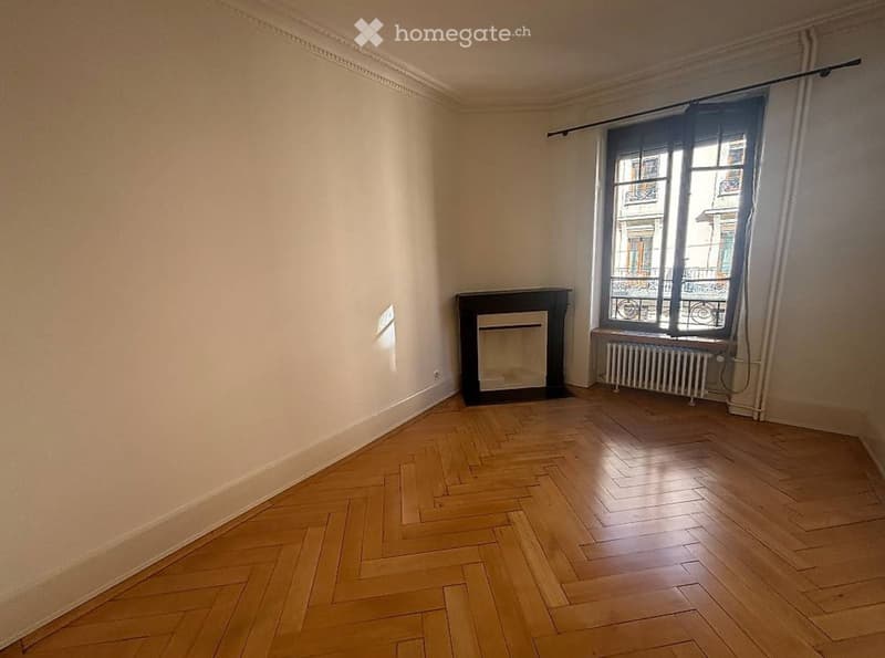 Empty room with wooden herringbone flooring, fireplace, large window with radiator below, and light-colored walls.