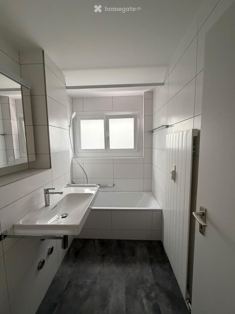 Modern bathroom with a white sink, bathtub, and large mirror. The room features tiled walls and a dark grey floor.