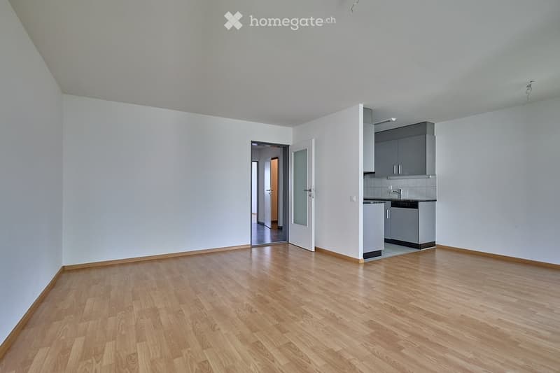 Empty room with wooden flooring, white walls, and a small kitchen area visible in the corner.
