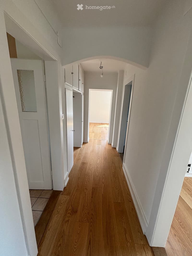 A long, narrow hallway with wooden flooring, white walls, and several doors leading to different rooms. The ceiling has a textured finish and a light fixture is visible.