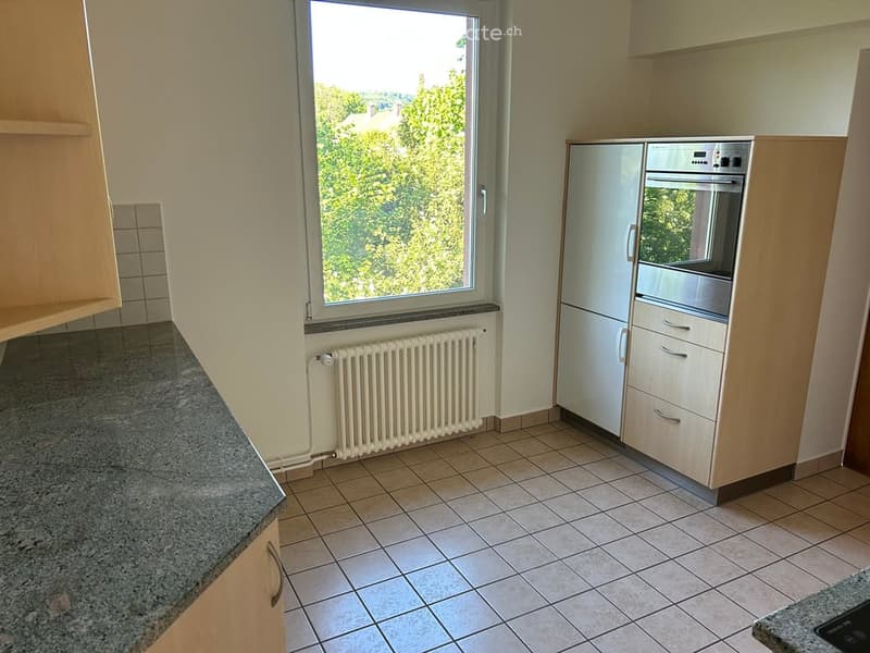 Modern kitchen with light wooden cabinets, granite countertops, tiled floor, and a large window offering a view of greenery.
