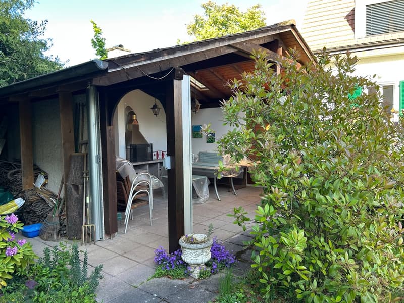 Wooden pergola-covered outdoor area with a stone floor, outdoor furniture, and surrounding greenery.