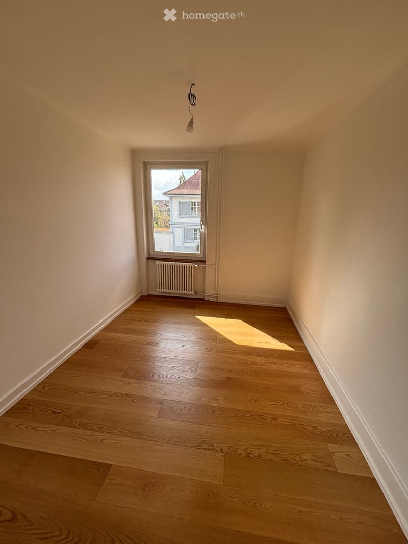 Empty room with wooden flooring, light-colored walls, a window with a radiator below, and a single light fixture hanging from the ceiling.