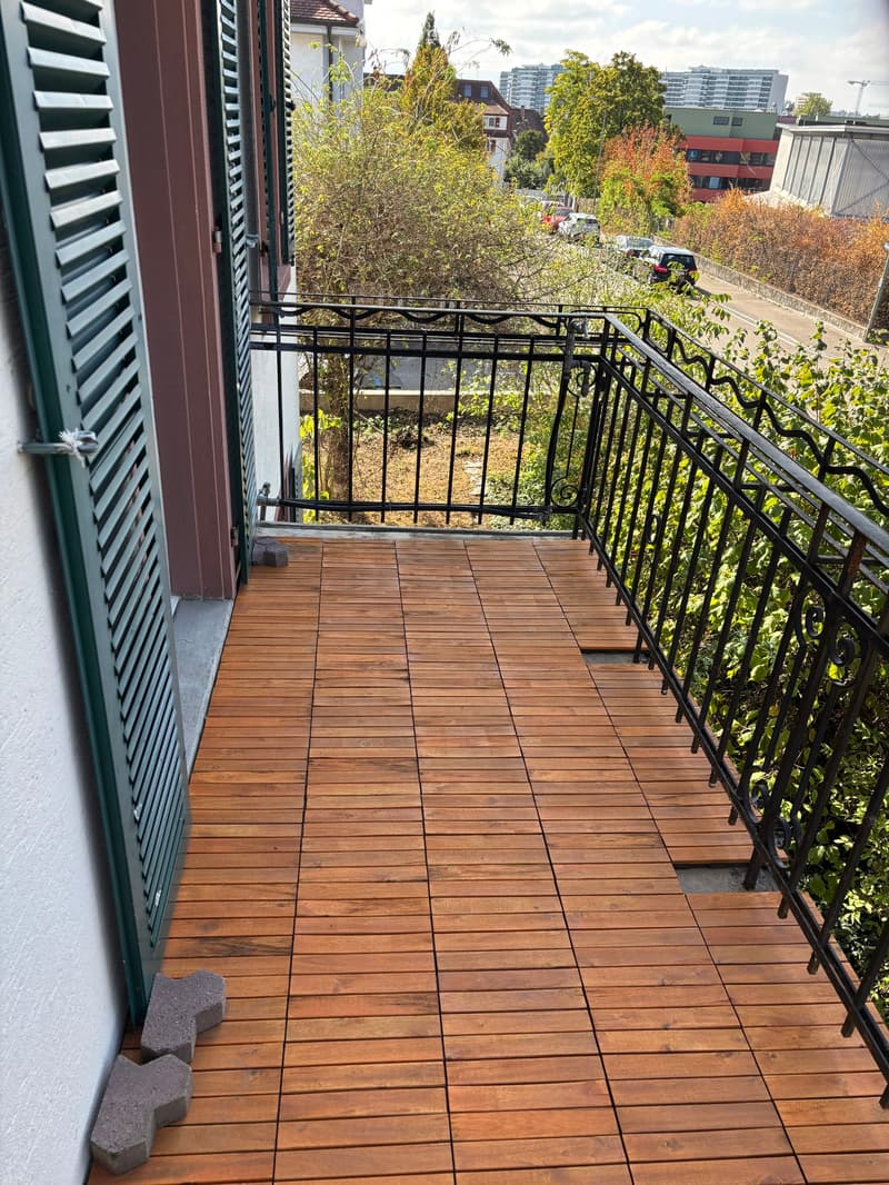 Wooden deck with black metal railing, overlooking a green area with trees and buildings in the background.