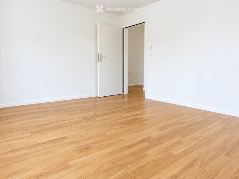 Empty room with light wooden flooring and white walls, featuring an open doorway leading to another room.