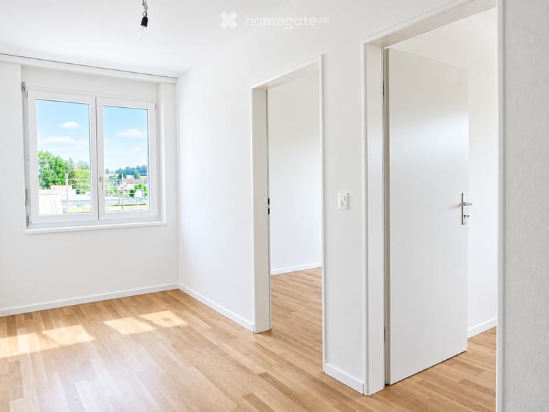 Empty room with white walls, light wooden flooring, and a large window with a view of greenery. A closed white door is visible on the right side.