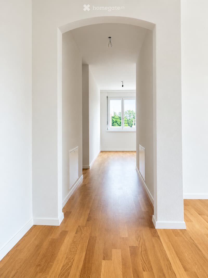 Empty hallway with light wooden flooring, white walls, and a large window at the end showing greenery outside.