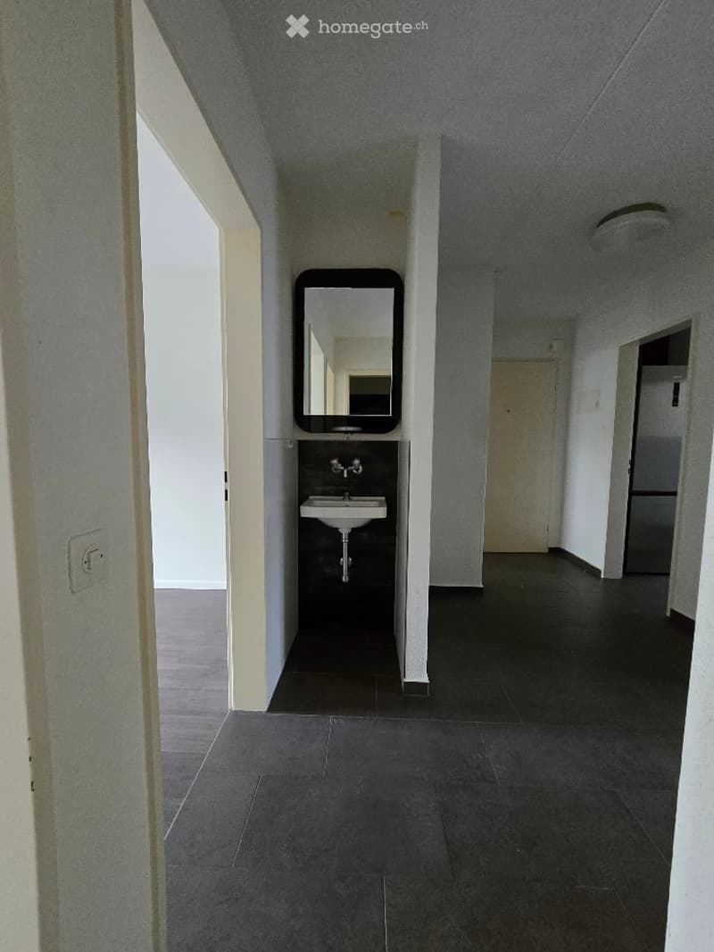 Empty hallway with a small sink and mirror in one corner, leading to other rooms.