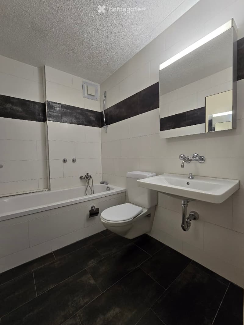 Modern bathroom with white tiles, black accents, bathtub, toilet, sink, and large mirror with built-in lighting.