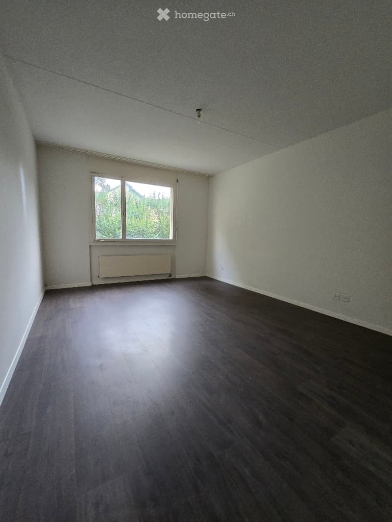 Empty room with wooden flooring, white walls, and a window with a radiator below it.