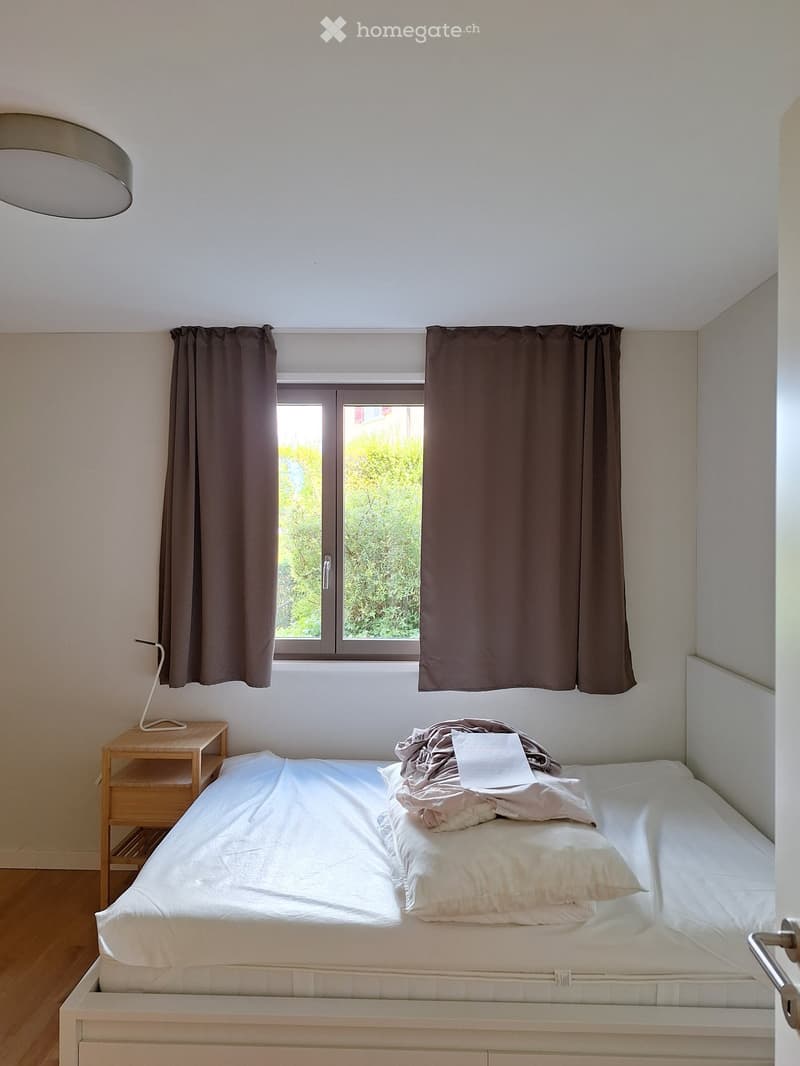Minimalist bedroom with a single bed, nightstand, lamp, and curtains. The bed is neatly made with white bedding and pillows.