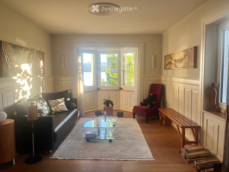 Bright living room with large windows, wooden flooring, a black leather sofa, red armchair, glass coffee table, wooden bench, and decorative items.