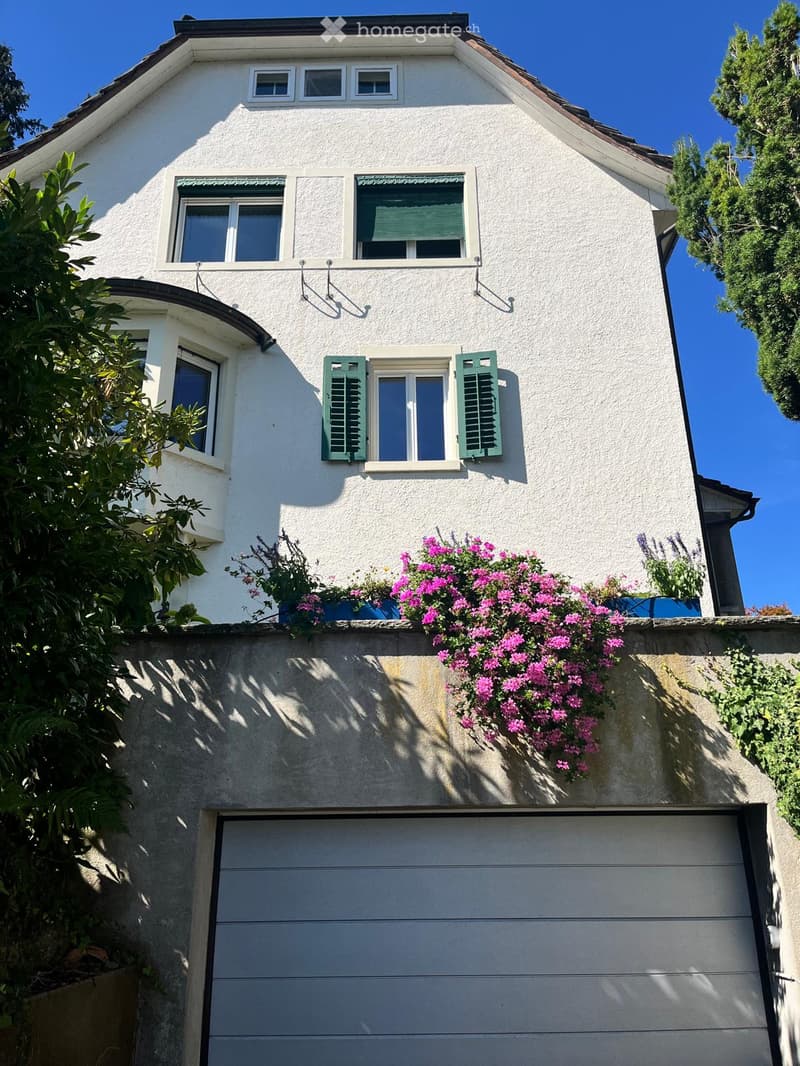 Two-story house with white exterior, green shutters, multiple windows, and a garage. Pink flowers adorn the roofline.