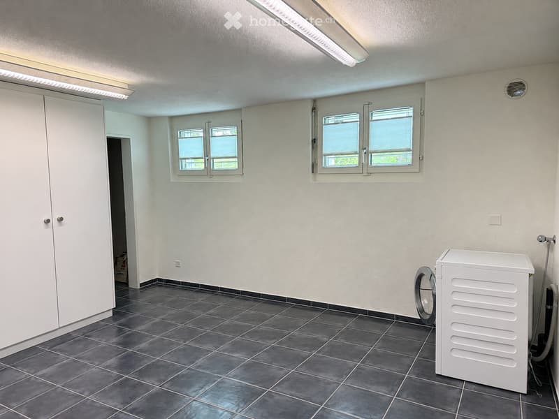 Empty room with tiled floor, white walls, and a private washing machine in the corner. Windows with blinds and built-in storage units are visible.
