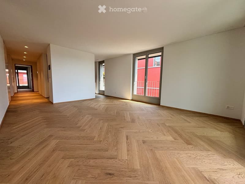 Empty hallway with wooden flooring, white walls, and recessed lighting. Features a large window with a view of a red building outside.