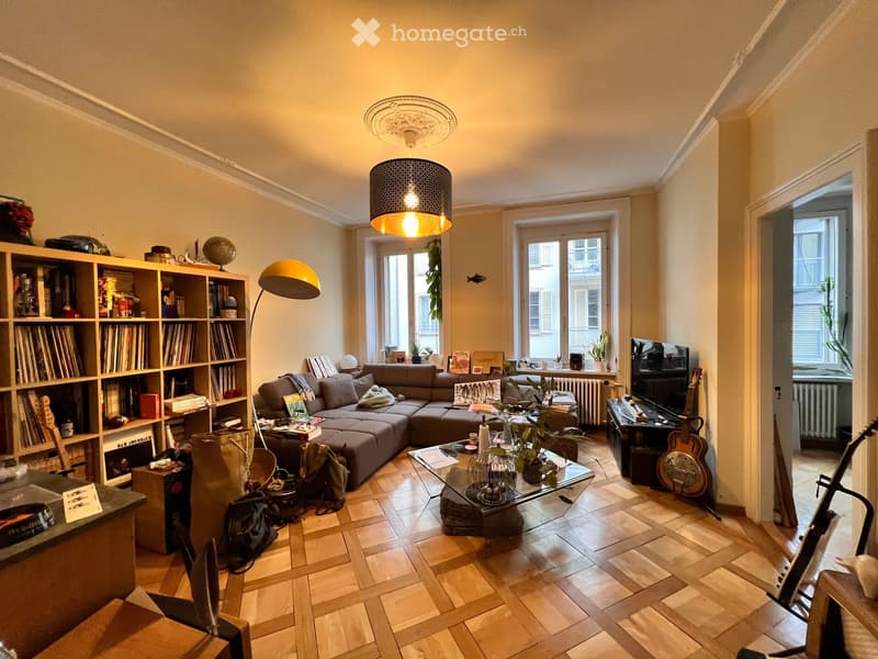 A cozy living room with a sectional sofa, coffee table, bookshelf, floor lamp, and guitar. The room features wooden flooring and large windows letting in natural light.