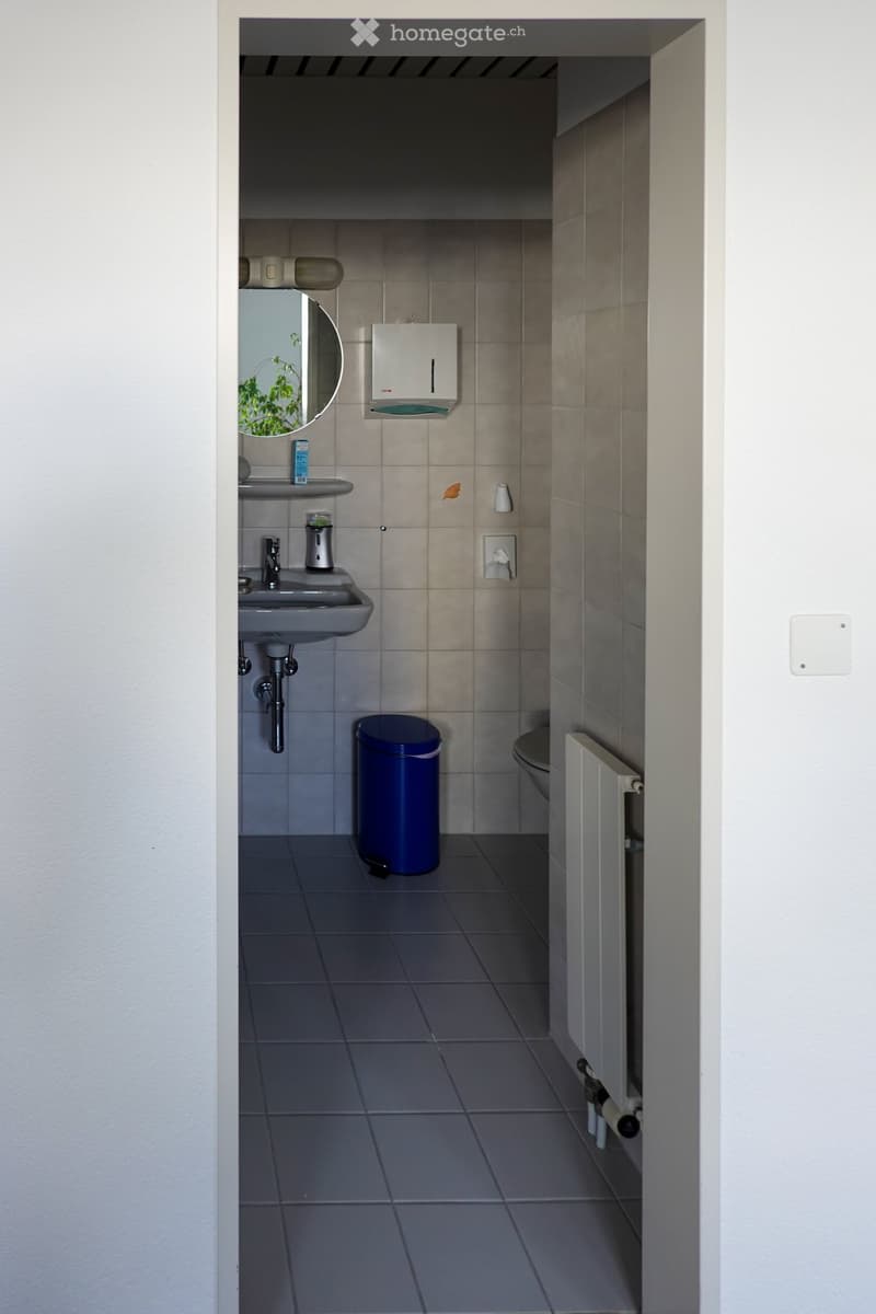 Modern bathroom with tiled walls and floor, featuring a sink with a mirror, a toilet, a blue trash can, and a hand dryer.