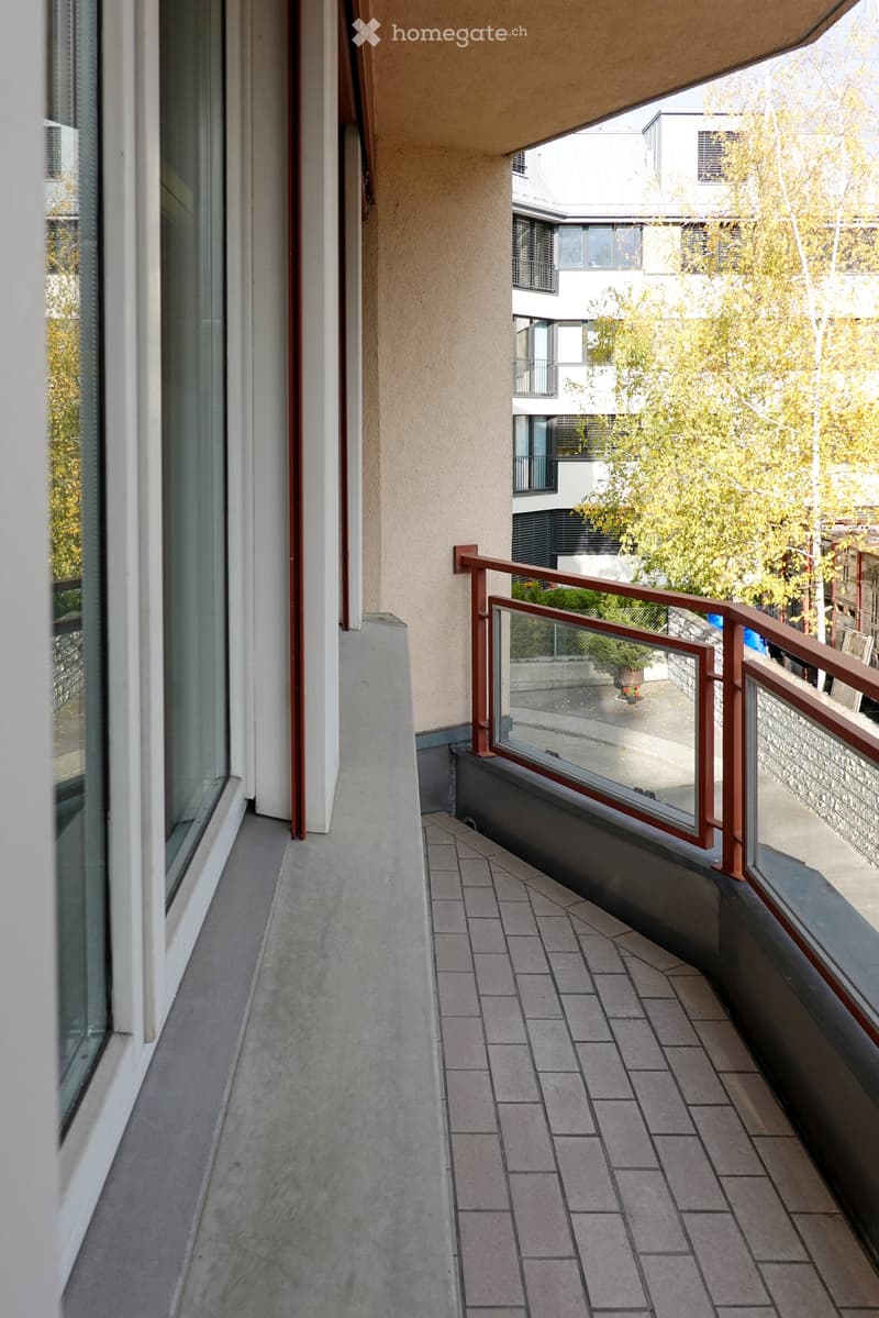 Balcony with tiled floor, metal railing, and glass panels, overlooking a residential area with trees and neighboring buildings.