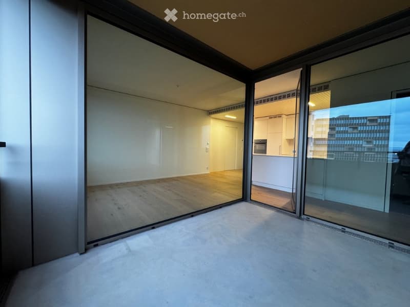 Empty room with large glass sliding doors, wooden flooring, and a view of a modern kitchen area in the background.