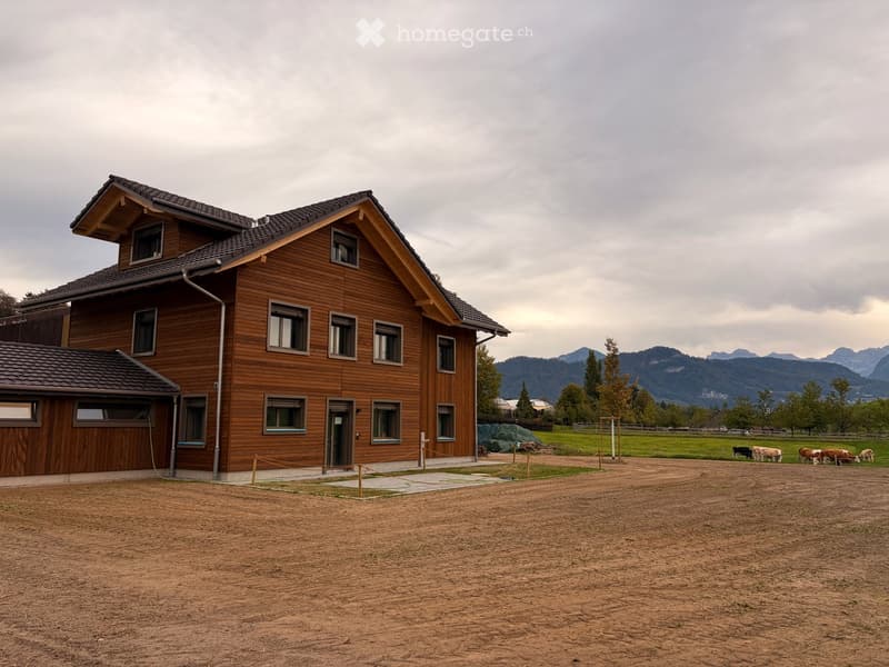 Two-story wooden house with multiple windows and a garage, surrounded by a grassy field with cows grazing in the background and mountains visible in the distance.
