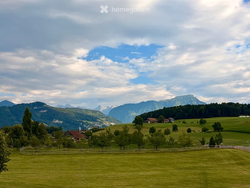 A serene rural landscape with green fields, scattered trees, a few houses, and distant mountains under a partly cloudy sky.