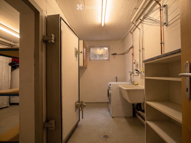 Utility room with a washing machine, sink, shelves, and exposed pipes.