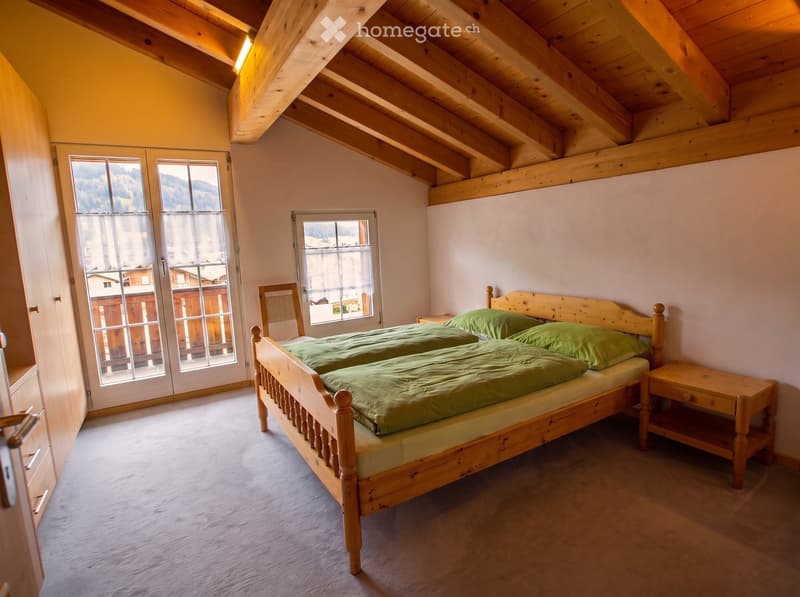 Cozy attic bedroom with wooden beams, large windows offering a scenic mountain view, a double bed with green bedding, a nightstand, and a wooden chair.