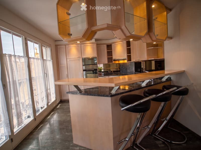 Modern kitchen with white cabinets, a central bar counter, and two black bar stools. Features large windows with white curtains and a sleek, contemporary design.