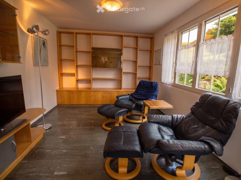 Modern living room with dark wooden flooring, large windows with white curtains, two leather chairs with matching ottomans, a small wooden side table, built-in wooden shelving unit, and a TV stand with a flat-screen TV.