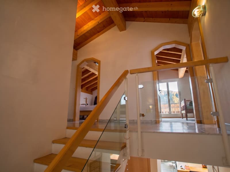 Staircase with wooden steps and glass railing, leading to a bedroom area with visible bed and furniture.