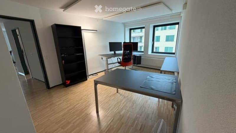 Bright, modern office space with wooden flooring, large windows, a desk with a computer, a bookshelf, and a comfortable chair.