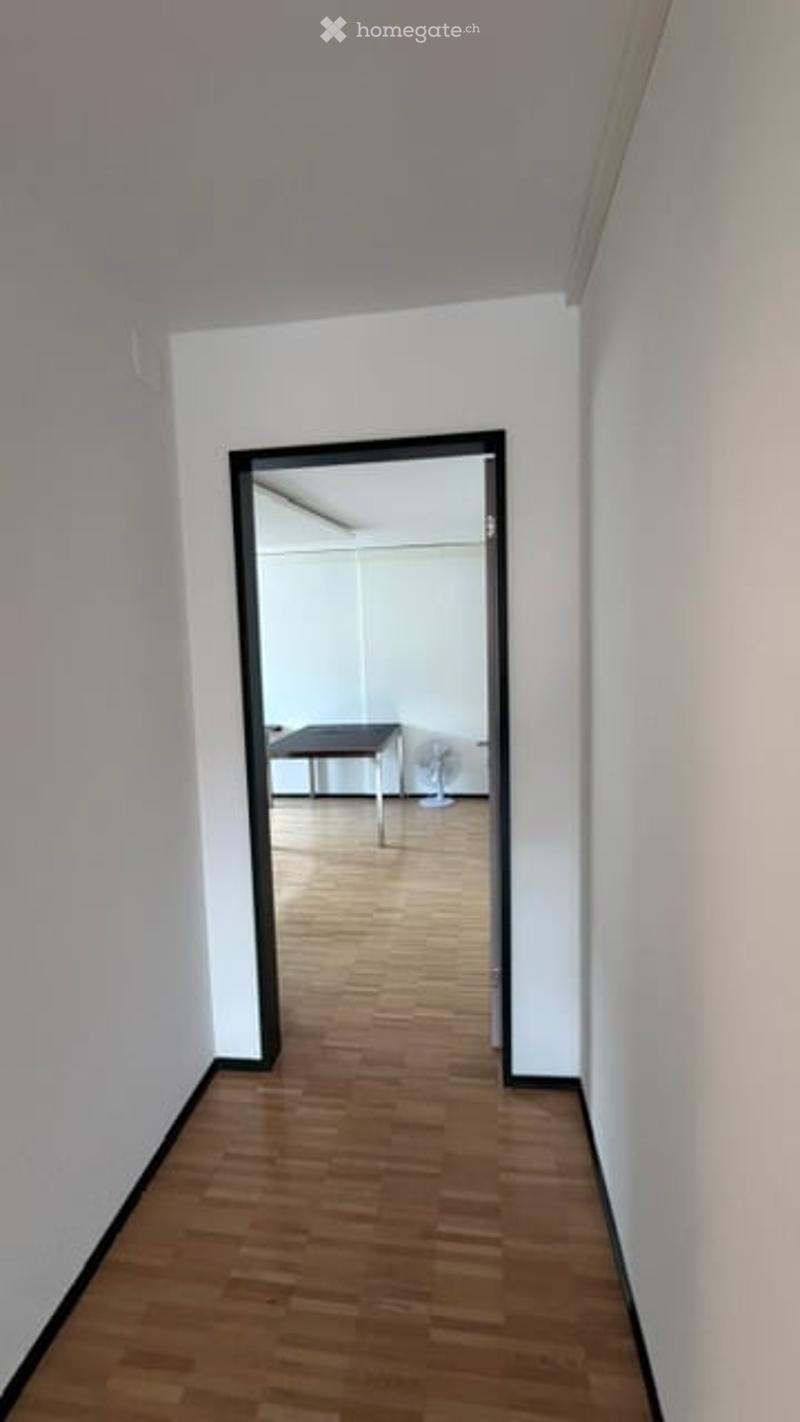 Empty hallway with wooden flooring and white walls, leading to an adjacent room with a table and fan.