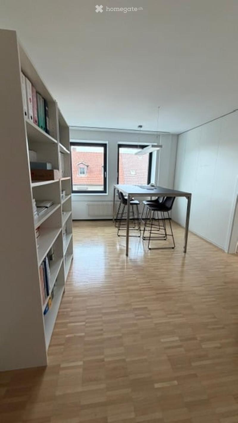 Bright, minimalist room with wooden flooring, large windows, bookshelf filled with books, and a small table with chairs.