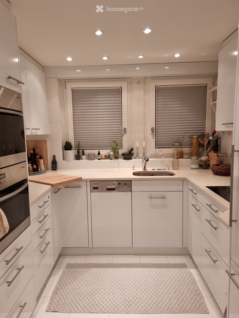 Modern white kitchen with built-in oven, dishwasher, sink, and various kitchen items on the counter, including jars, a cutting board, and small decorative items.