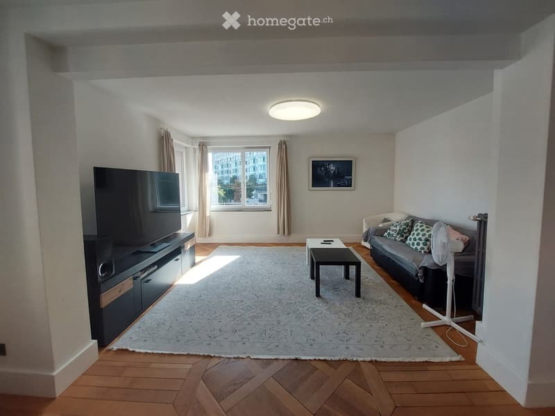 Modern living room with wooden flooring, large window, grey sofa, coffee table, TV, and fan.