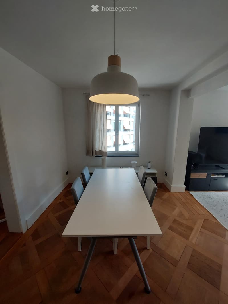 Modern dining area with a long white dining table, four chairs, a pendant light fixture, and a television on a stand. The room features wooden flooring and large windows with curtains.
