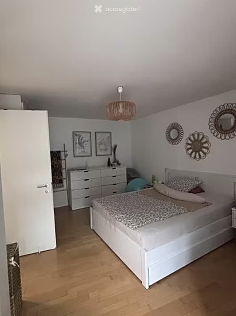 Modern bedroom with a white bed, nightstand, dresser, and decorative wall mirrors. The room features light wooden flooring and a hanging woven lamp.