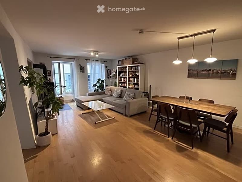 Open-plan living and dining area with wooden flooring, a grey sectional sofa, a glass coffee table, a wooden dining table with chairs, a bookshelf, and a potted plant near the window.