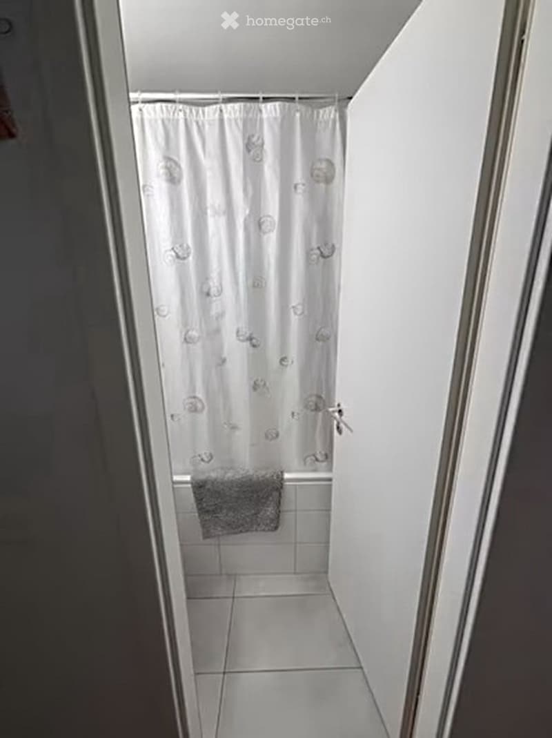 Small bathroom with white tiled walls and floor, a shower curtain with a floral pattern, and a grey towel hanging on the side.