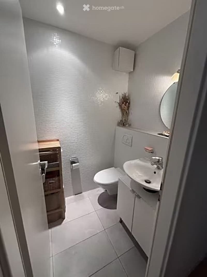 Modern, compact bathroom with white tiled walls and floor, a toilet, a sink with a mirror, and a small cabinet.