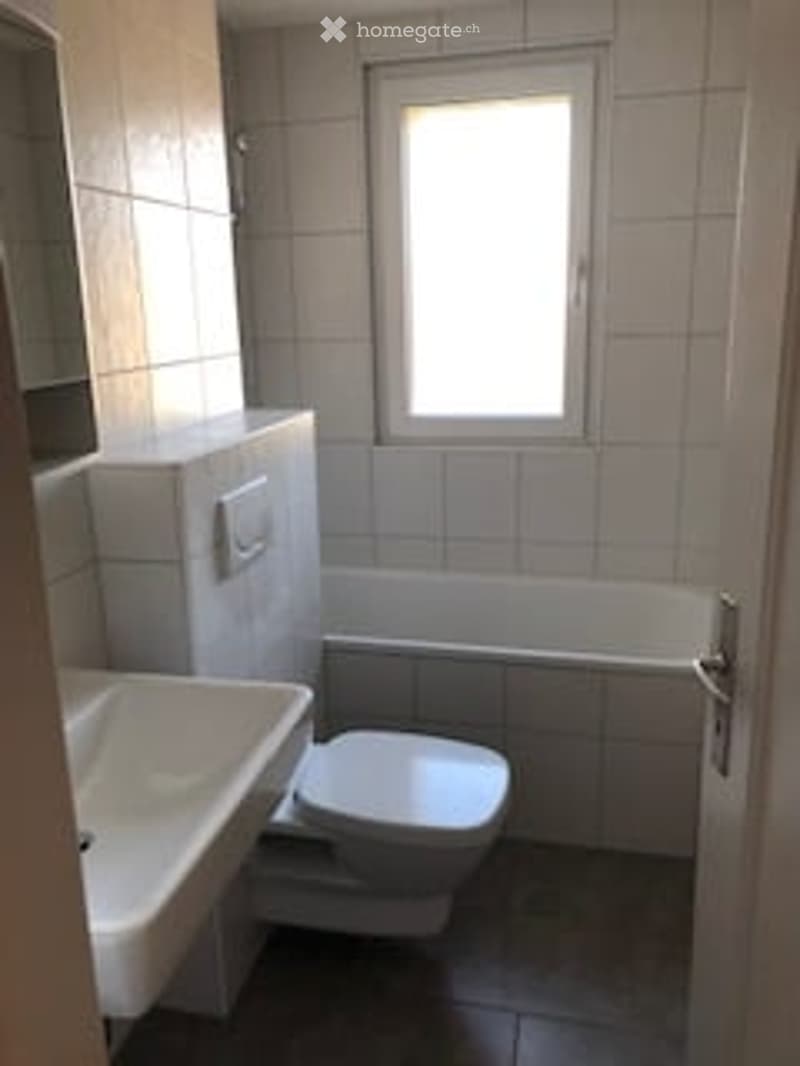 A small bathroom with white tiles, a toilet, a sink, and a bathtub. The room has a window for natural light and a door for access.